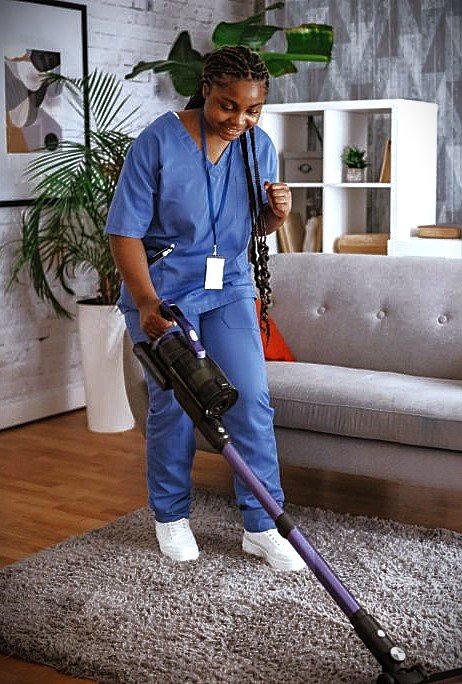 CareNestPro_A person in blue scrubs is vacuuming a carpet in a stylish living area featuring modern furniture and vibrant home decor items, maintaining a clean environment.